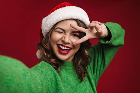 Photo of happy young woman in christmas santa hat taking a selfie by camera isolated over red wall backgroundの写真素材