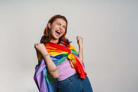 Excited woman making winner gesture while posing with rainbow flag isolated over gray backgroundの写真素材
