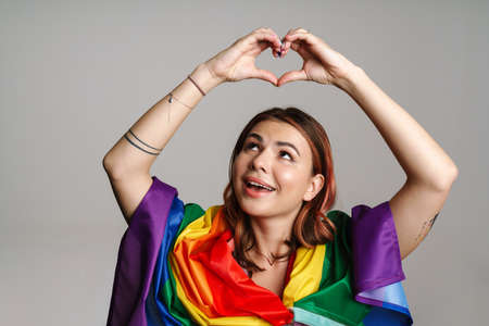 Cheerful woman wearing rainbow flag smiling and showing heart gesture isolated over gray backgroundの写真素材
