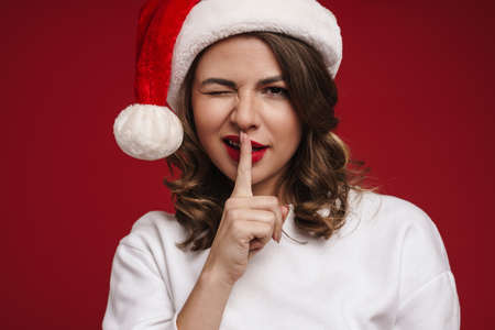 Photo of positive cheery young woman in christmas santa hat isolated over red wall background showing silence gestureの写真素材