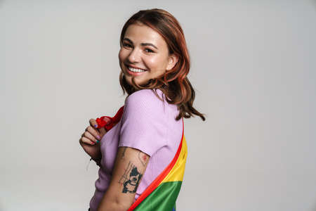 Happy beautiful woman smiling while posing with rainbow flag isolated over gray backgroundの写真素材