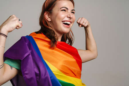 Cheerful woman wearing rainbow flag laughing and showing her biceps isolated over gray backgroundの写真素材