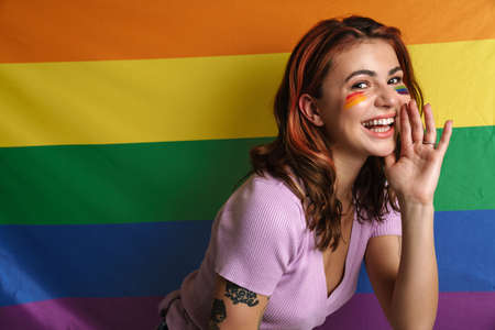 Cheerful young woman with makeup whispering secret isolated over rainbow flagの写真素材