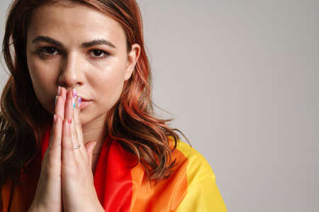 Focused beautiful young woman wearing rainbow flag holding palms together isolated over gray backgroundの写真素材