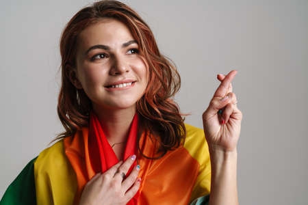 Cheerful woman wearing rainbow flag holding fingers crossed for good luck isolated over gray backgroundの写真素材