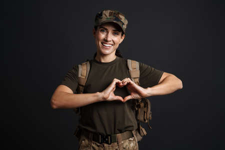 Smiling soldier woman with army backpack showing heart gesture isolated over black backgroundの写真素材