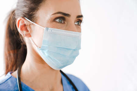 Beautiful young woman doctor in medical mask and uniform isolated over white background, close up