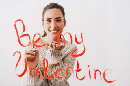 Beautiful smiling young woman in love drawing be my valentine sign in front of her isolated over...