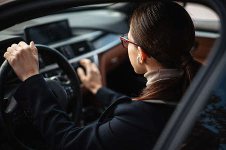 Beautiful focused businesswoman in earphones and eyeglasses driving car