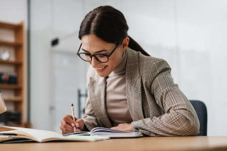 Happy charming woman smiling and writing down notes in planner while working at officeの写真素材