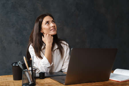 Smiling mid aged brunette businesswoman in shirt sitting at the desk with laptop computer over dark...