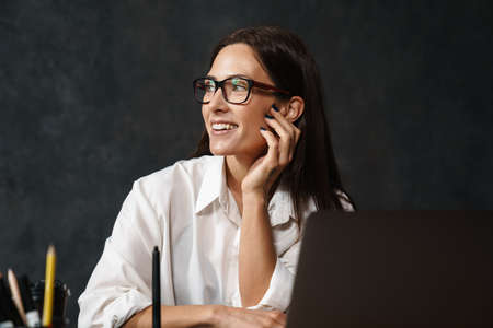 Smiling mid aged brunette businesswoman in shirt sitting at the desk with laptop computer over dark...