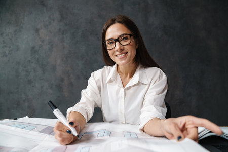 Smiling mid aged business woman in white shirt sitting at the desk with paperwork over dark wall backgroundの写真素材