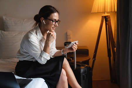 Smiling mid aged brunette white businesswoman sitting on bed in a hotel room on a video call via mobile phone and wireless earphonesの写真素材