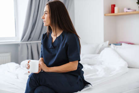 A brunette woman in a blue medical uniform sitting on the bed and holding a white cup in her hands while looking to the side against the background of the roomの写真素材