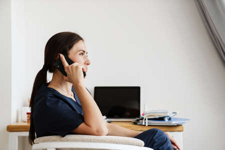 The side view of medical woman in medical uniform sitting on chair at table and talking on phone in...