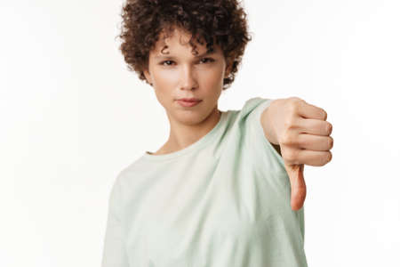 Young curly brunette woman showing thumb down at camera isolated over white background