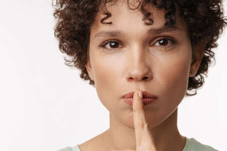 Young curly woman looking at camera and showing silence gesture isolated over white backgroundの写真素材