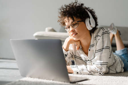 Young curly woman in headphones using laptop while lying on floor at home