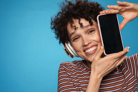 Young curly smiling woman wearing headphones showing cellphone isolated blue white backgroundの写真素材