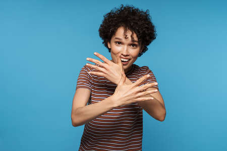 Young curly brunette woman gesturing and smiling at camera isolated over blue background