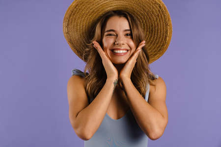 Young white woman in straw hat smiling and grabbing her face isolated over purple background