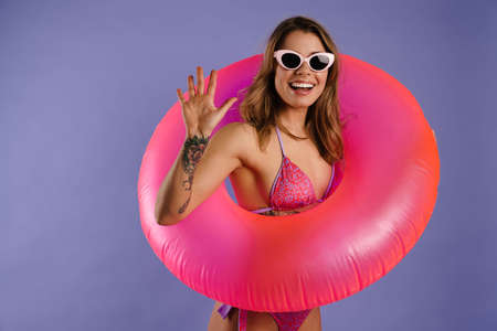 Young white woman in swimsuit smiling and holding rubber ring isolated over purple background
