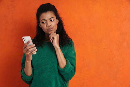 Pensive african casual brunette woman standing over orange wall background, looking at mobile phone...