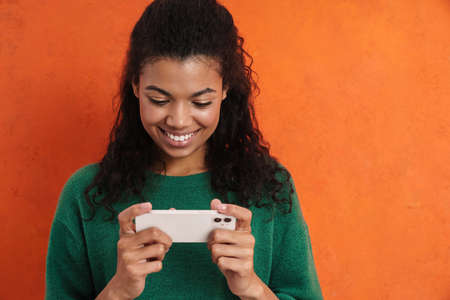 Happy smiling african casual brunette woman standing over orange wall background, holding mobile...