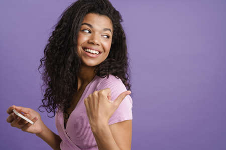 Young black woman pointing finger aside and using mobile phone isolated over purple background