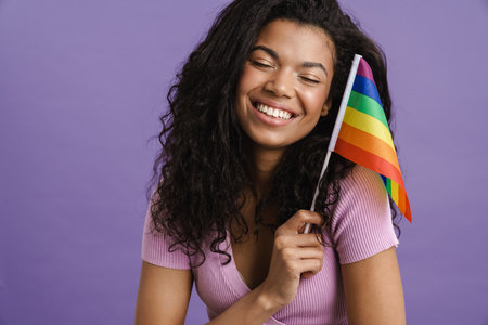 Young black woman smiling while posing with rainbow flag isolated over purple backgroundの写真素材
