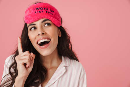 Happy beautiful woman smiling while posing in sleeping mask isolated over pink background