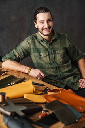 Bearded happy craftsman working with leather while sitting at table in workshop