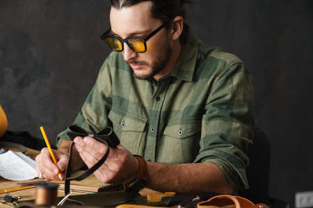 Bearded focused craftsman working with leather while sitting at table in workshop