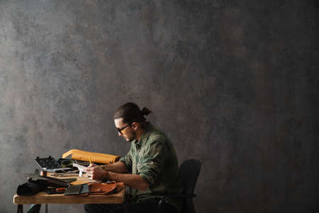 Bearded focused craftsman writing down notes while working with leather in workshopの写真素材