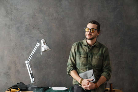 Bearded white craftsman standing with leather clutch in his workshop