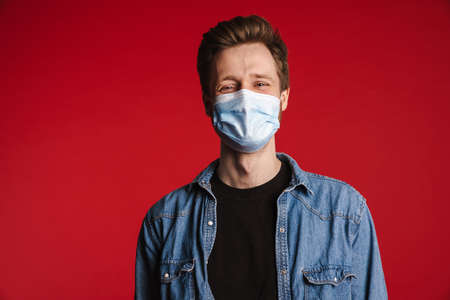 Young man in casual clothes wearing protective medical mask standing over red wall background