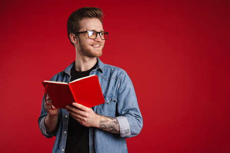 Happy smiling young man in casual clothes standing over red wall background, reading a book, holding pencilの写真素材