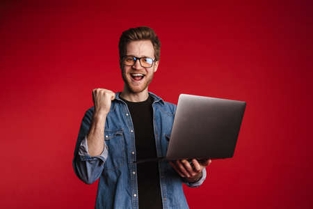 Happy smiling young man in casual clothes standing over red wall background, holding laptop...