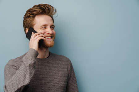 White ginger man with beard smiling while talking on mobile phone isolated over blue background