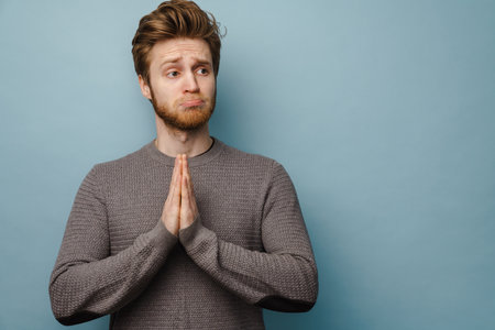 White ginger man grimacing while holding palms together isolated over blue backgroundの写真素材