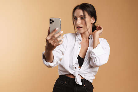 Young brunette woman in shirt taking selfie on cellphone isolated over beige background