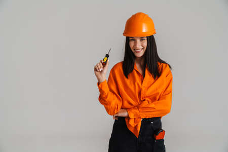 Young woman in helmet smiling while posing with screwdriver isolated over grey background