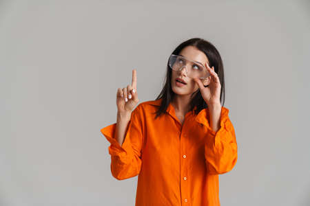 Young brunette woman in safety glasses looking and pointing finger upward isolated over grey backgroundの写真素材