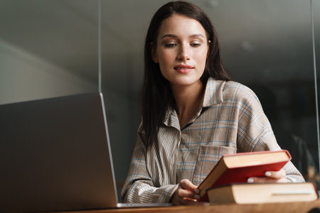 Young brunette woman reading book and working with laptop in officeの写真素材