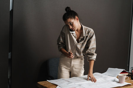 Young brunette woman talking on cellphone while working with drawings in officeの写真素材