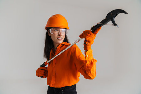 Young woman in safety glasses and helmet posing with building tool isolated over grey background