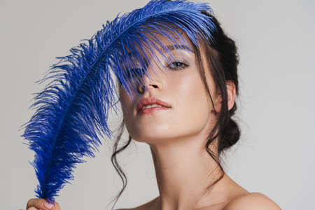 Young topless white woman with brown hair pulled up posing holding blue feather over gray background, close upの写真素材