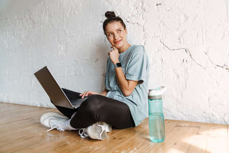 Smiling young white brunette woman in active wear sitting on a floor indoors with laptop computer...