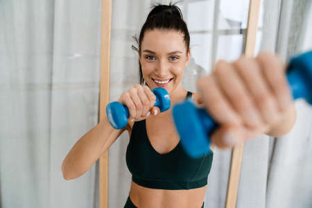 Happy brunette white woman lifting dumbbells at home in the living room
